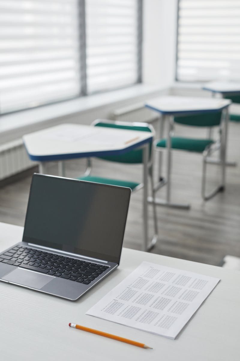 A laptop on a desk in a bright classroom with papers and a pencil nearby.